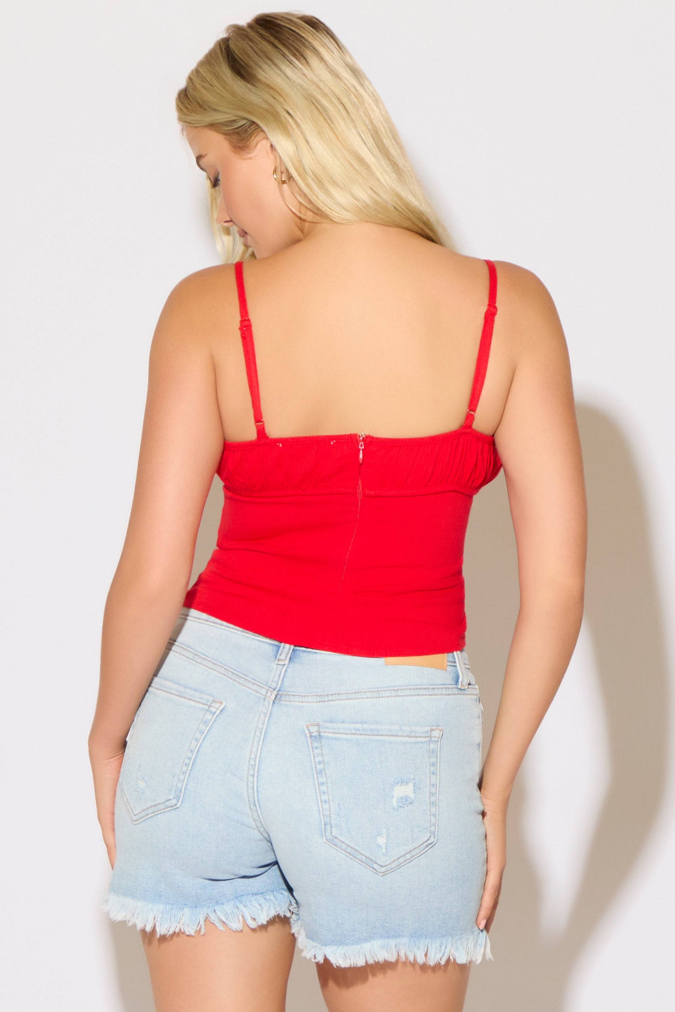 Model is wearing a red ruched bust strappy cami top with denim shorts on a white background. 