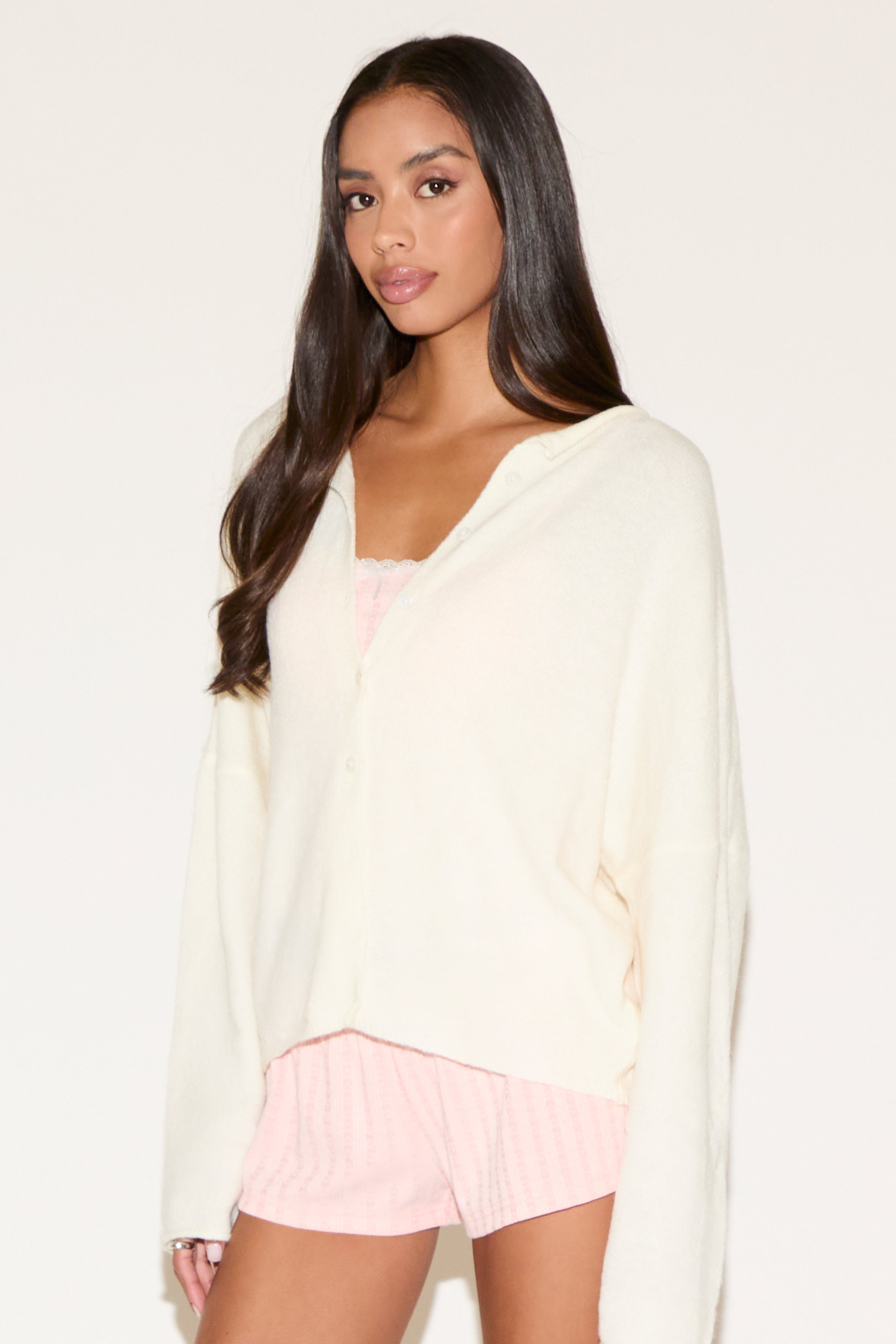 Woman wearing a white cardigan over a pink top and shorts on a plain background