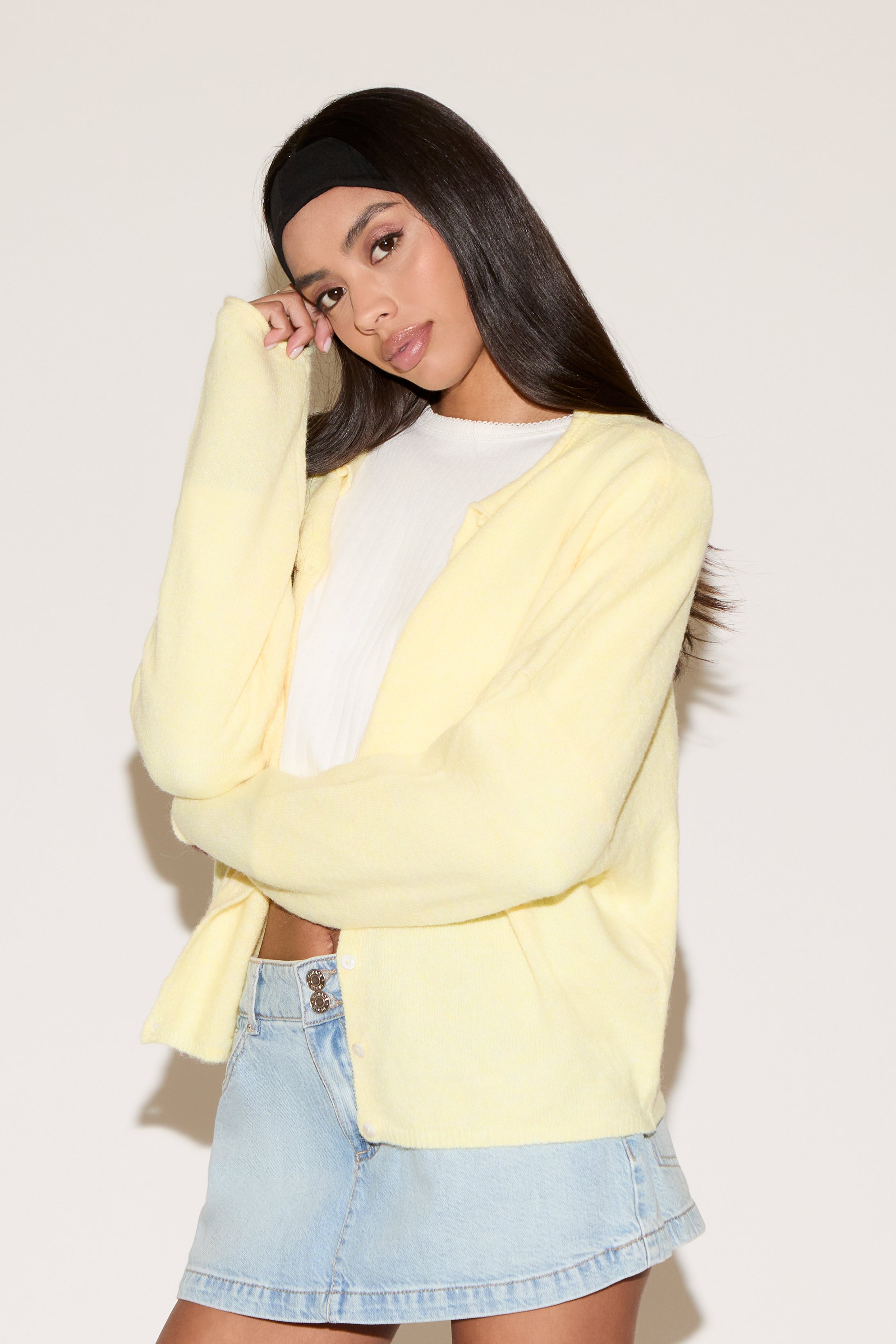Woman wearing a light yellow cardigan over a white top and denim skirt on a plain background
