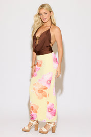 Model is wearing a brown tie neck and back metal detail cowled top with a yellow maxi skirt with pink and orange floral print on a white background.