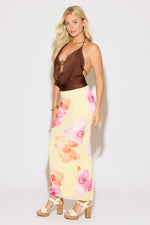 Model is wearing a brown tie neck and back metal detail cowled top with a yellow maxi skirt with pink and orange floral print on a white background.