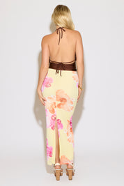 Model is wearing a brown tie neck and back metal detail cowled top with a yellow maxi skirt with pink and orange floral print on a white background.