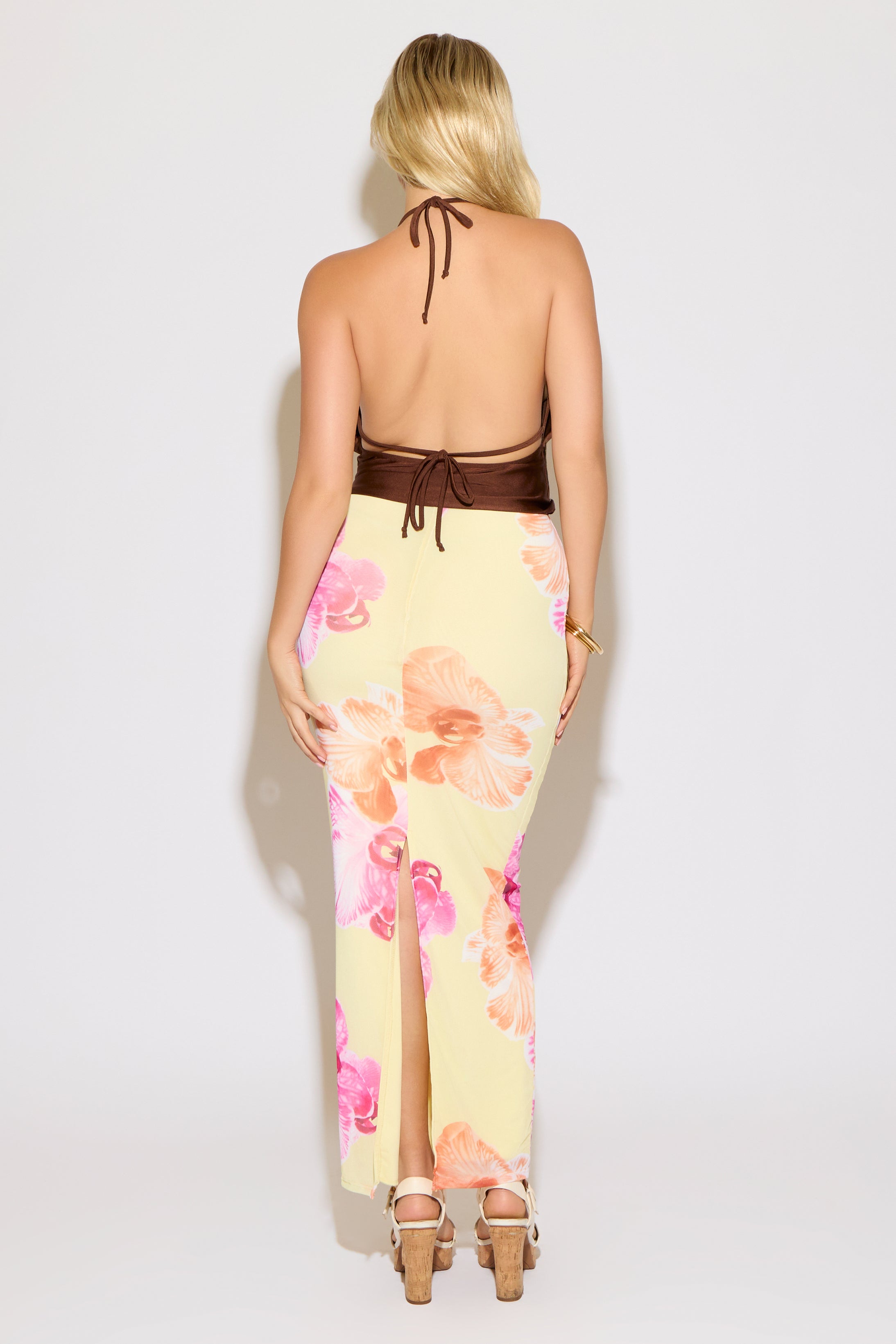 Model is wearing a brown tie neck and back metal detail cowled top with a yellow maxi skirt with pink and orange floral print on a white background.