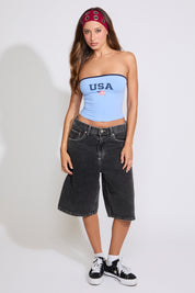 Model is wearing a blue USA strapless tube top with washed black denim jorts on a white background. 