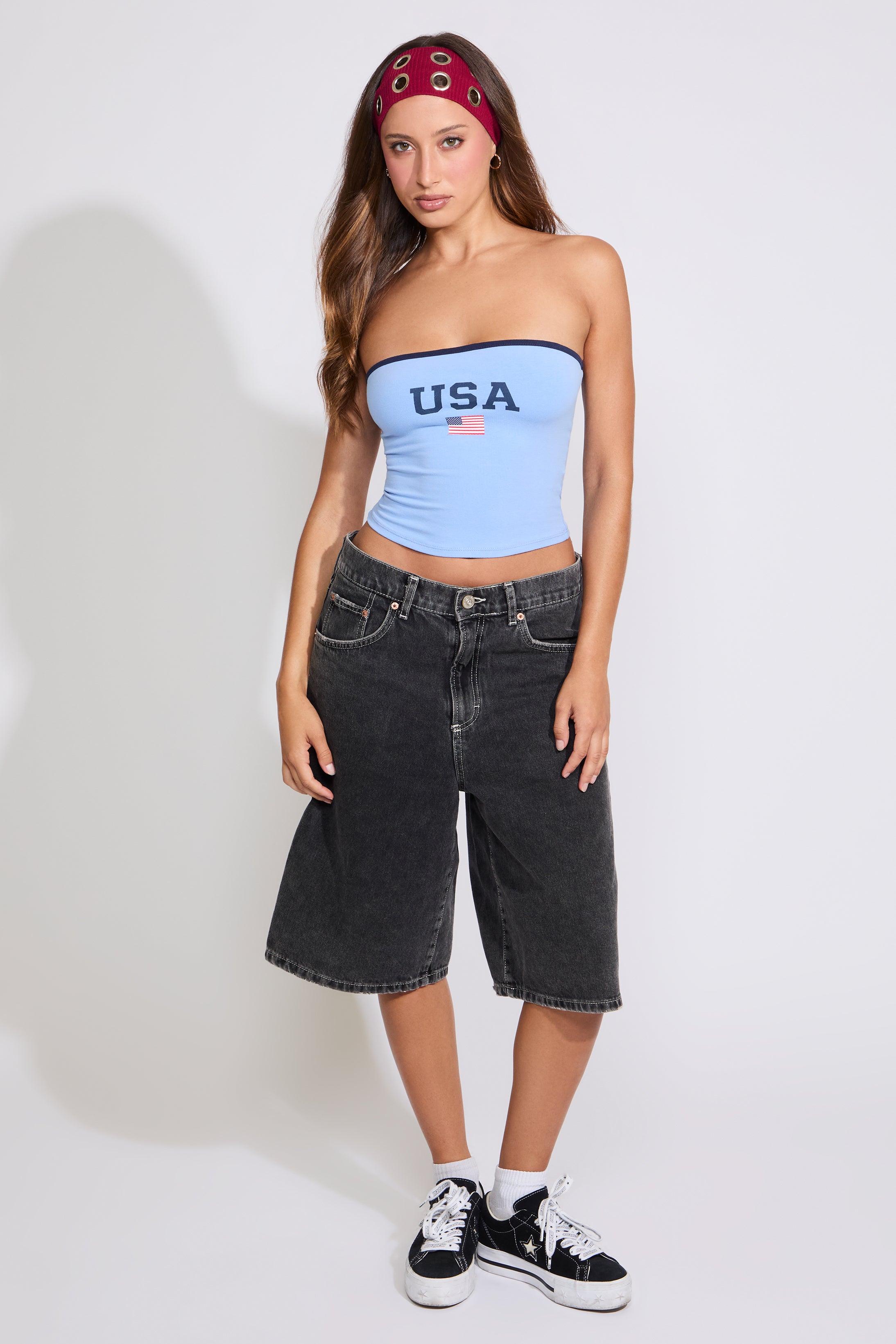 Model is wearing a blue USA strapless tube top with washed black denim jorts on a white background. 