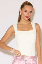 A woman stands against a white background wearing a cream sleeveless top paired with red-and-white striped knee-length pants and clear heeled sandals.