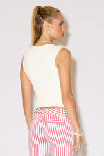 A woman stands against a white background wearing a cream sleeveless top paired with red-and-white striped knee-length pants and clear heeled sandals.
