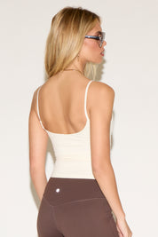 A woman wearing large sunglasses, a white tank top, and brown leggings stands against a plain, light background.