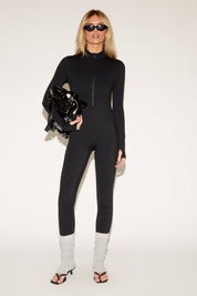 Woman in a black zip-front jumpsuit, white leg warmers, black high-heeled sandals, sunglasses, and holding a black jacket, poses against a plain background.