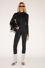 Woman in a black zip-front jumpsuit, white leg warmers, black high-heeled sandals, sunglasses, and holding a black jacket, poses against a plain background.