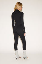 Woman in a black zip-front jumpsuit, white leg warmers, black high-heeled sandals, sunglasses, and holding a black jacket, poses against a plain background.