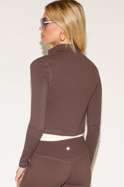 A woman wearing oversized sunglasses. She wears a brown zip-up jacket and matching leggings over a white top. 