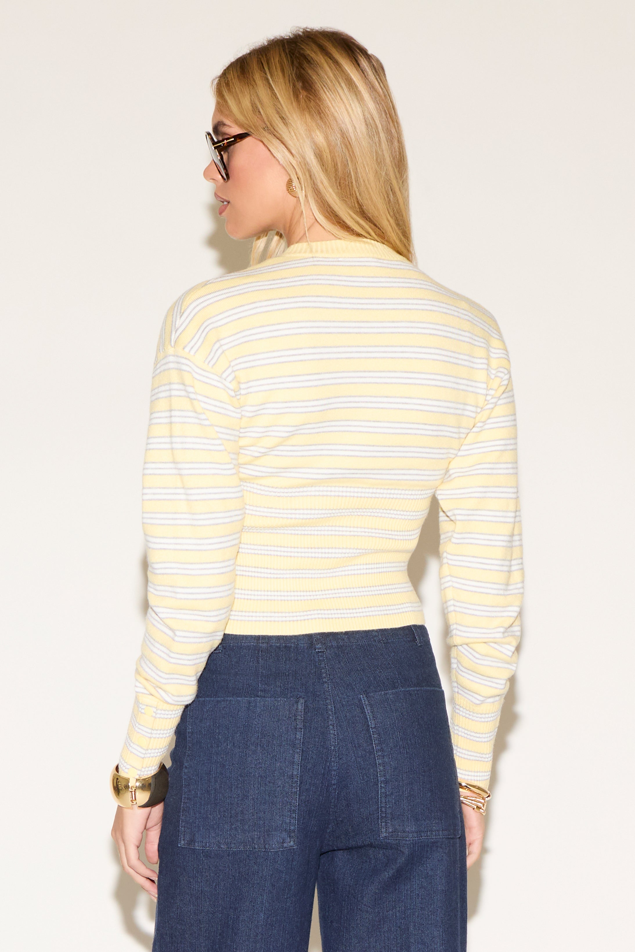 A woman in sunglasses wearing a yellow striped sweater and high-waisted wide-leg jeans poses confidently against a plain white background.