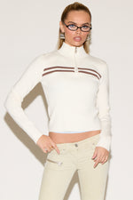 A person poses confidently in a white sweater with brown stripes, wearing glasses and beige pants. The expression is poised and stylish, set against a plain background.