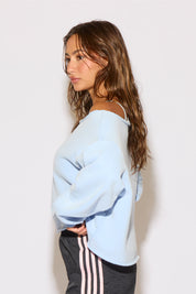 Woman wearing a light blue off-shoulder sweater and dark pants with white stripes against a plain background