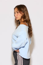 Woman wearing a light blue off-shoulder sweater and dark pants with white stripes against a plain background