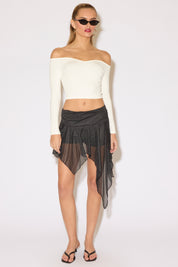 Woman in a chic off-the-shoulder white top and a black polka dot asymmetrical skirt stands confidently. She wears sunglasses and sandals, exuding a trendy vibe.