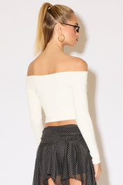 Woman in a chic off-the-shoulder white top and a black polka dot asymmetrical skirt stands confidently. She wears sunglasses and sandals, exuding a trendy vibe.