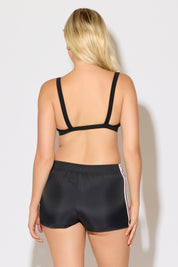 Model is wearing a black rib knit strap back cami bra top with black shorts on a white background. 