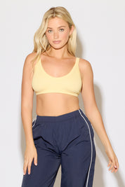 Model is wearing a butter yellow rib knit strap back cami bra top with navy blue pants on a white background. 