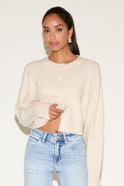 Model wearing a cream cropped button-up cardigan with a relaxed fit, styled with light-wash distressed denim jeans and gold statement earrings, posed against a neutral background.