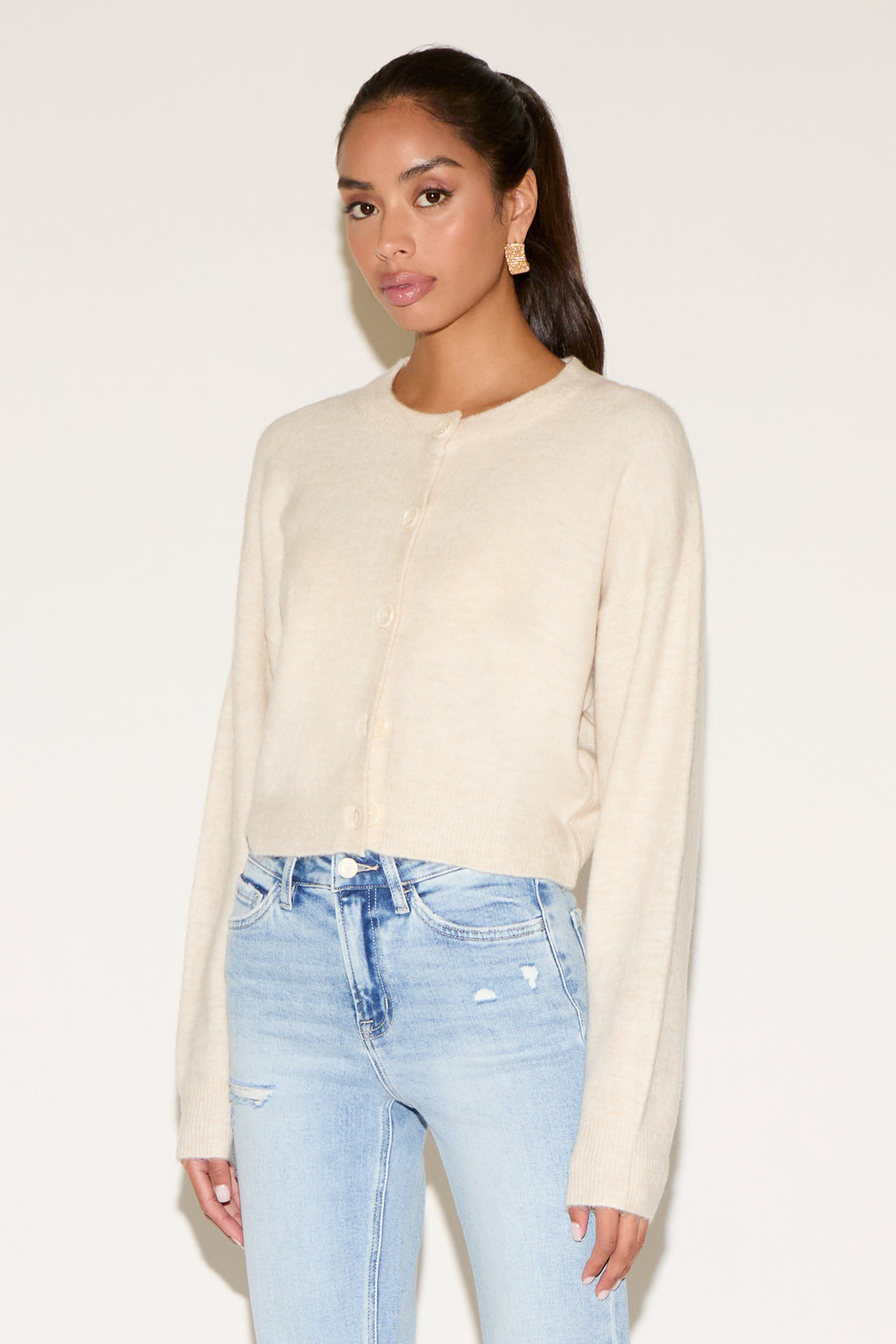 Model wearing a light gray button-up cardigan with a relaxed fit, styled with light-wash denim jeans, oversized tortoiseshell sunglasses, and gold statement earrings against a neutral background.
