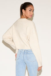 Back view of model wearing a cream cropped cardigan with a relaxed fit, styled with light-wash denim jeans and gold statement earrings, hair pulled into a sleek ponytail against a neutral background.