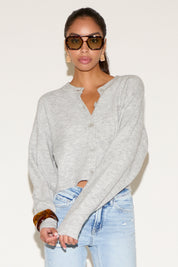 Model wearing a light gray button-up cardigan with a relaxed fit, styled with light-wash denim jeans, oversized tortoiseshell sunglasses, and gold statement earrings against a neutral background.