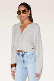 Model wearing a light gray cropped button-up cardigan with a relaxed fit, styled with light-wash denim jeans, tortoiseshell sunglasses, gold statement earrings, and stacked bangles against a neutral background.