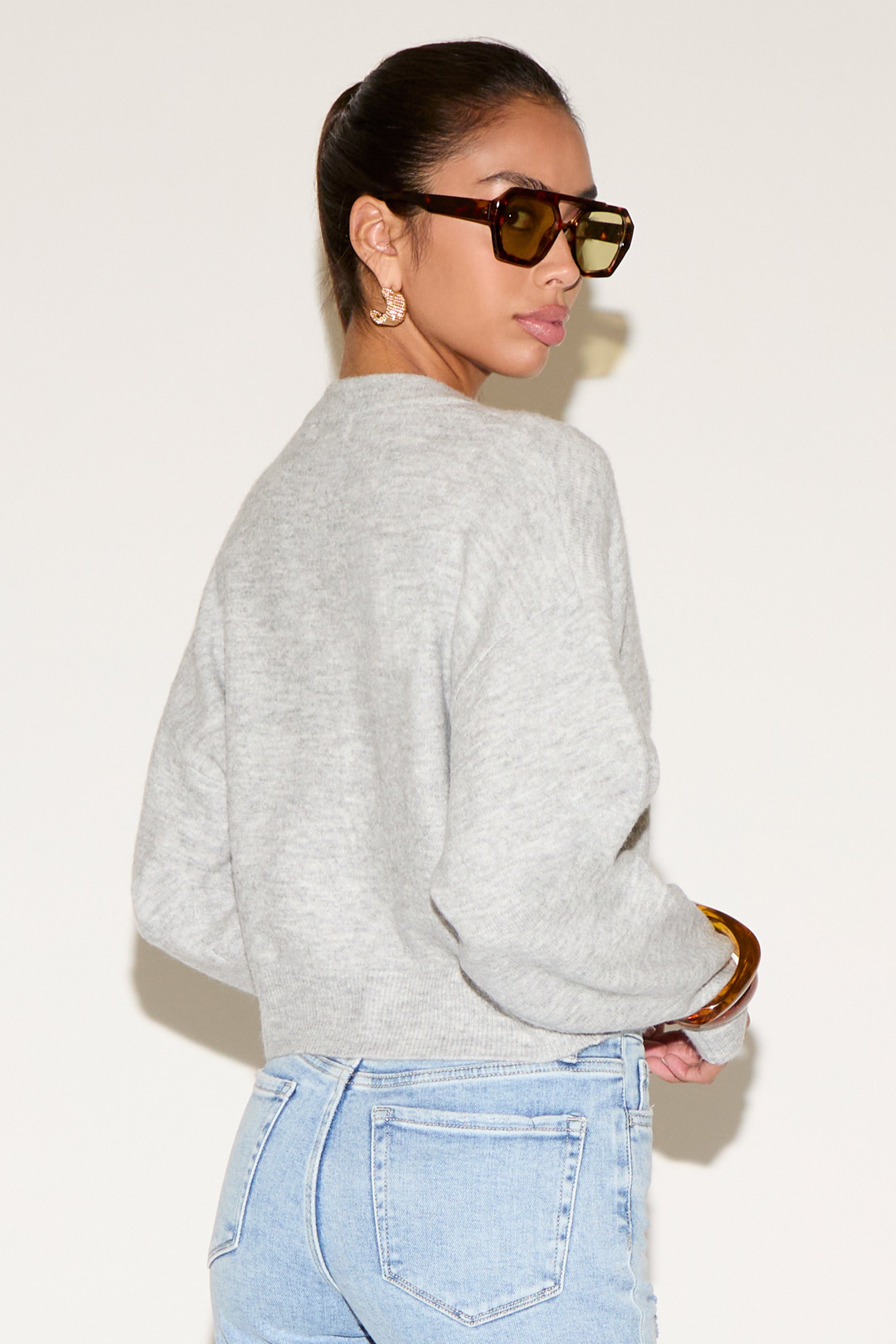 Back view of model wearing a light gray button-up cardigan with a relaxed fit, styled with light-wash denim jeans, tortoiseshell sunglasses, and gold statement earrings against a neutral background.