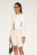Woman wearing a white cardigan and beige shorts with a plain background