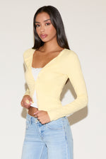 Woman wearing a light yellow cardigan over a white top and blue jeans on a plain background