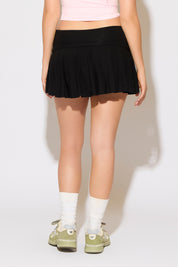 Model is wearing a black fold over waist lettuce hem ruffle mini skirt with a pink top on a white background.