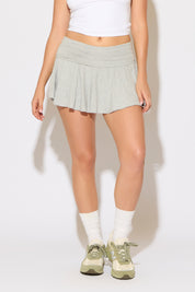 Model is wearing a heather grey fold over waist lettuce hem ruffle mini skirt with a white top on a white background.