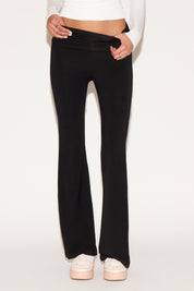 Model wearing black low-rise flare pants with a fold-over waistband, styled with a white long-sleeve top and white sneakers against a neutral background.