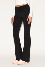 Model wearing black low-rise flare pants with a fold-over waistband, styled with a white long-sleeve top and white sneakers against a neutral background.