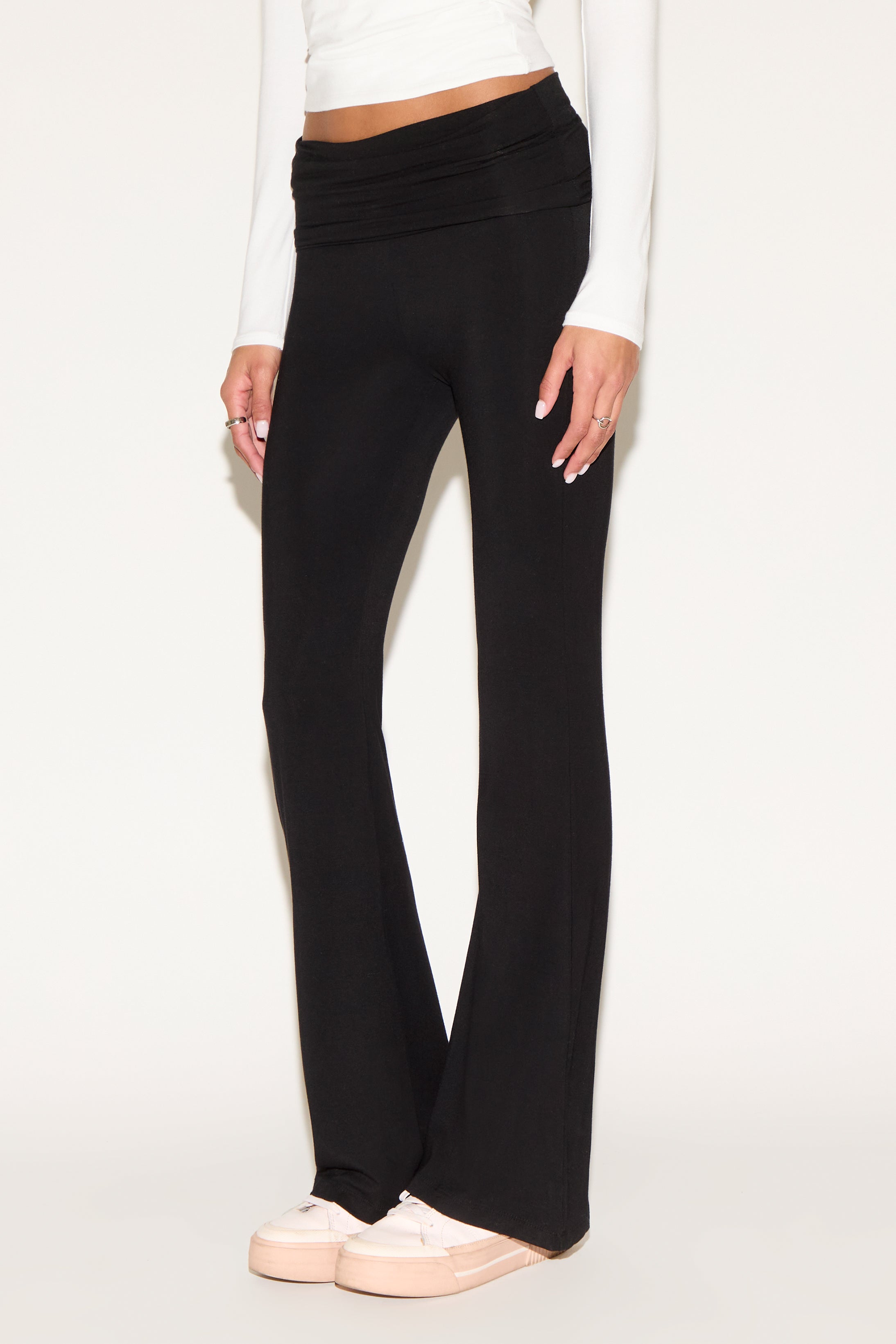 Model wearing black low-rise flare pants with a fold-over waistband, styled with a white long-sleeve top and white sneakers against a neutral background.