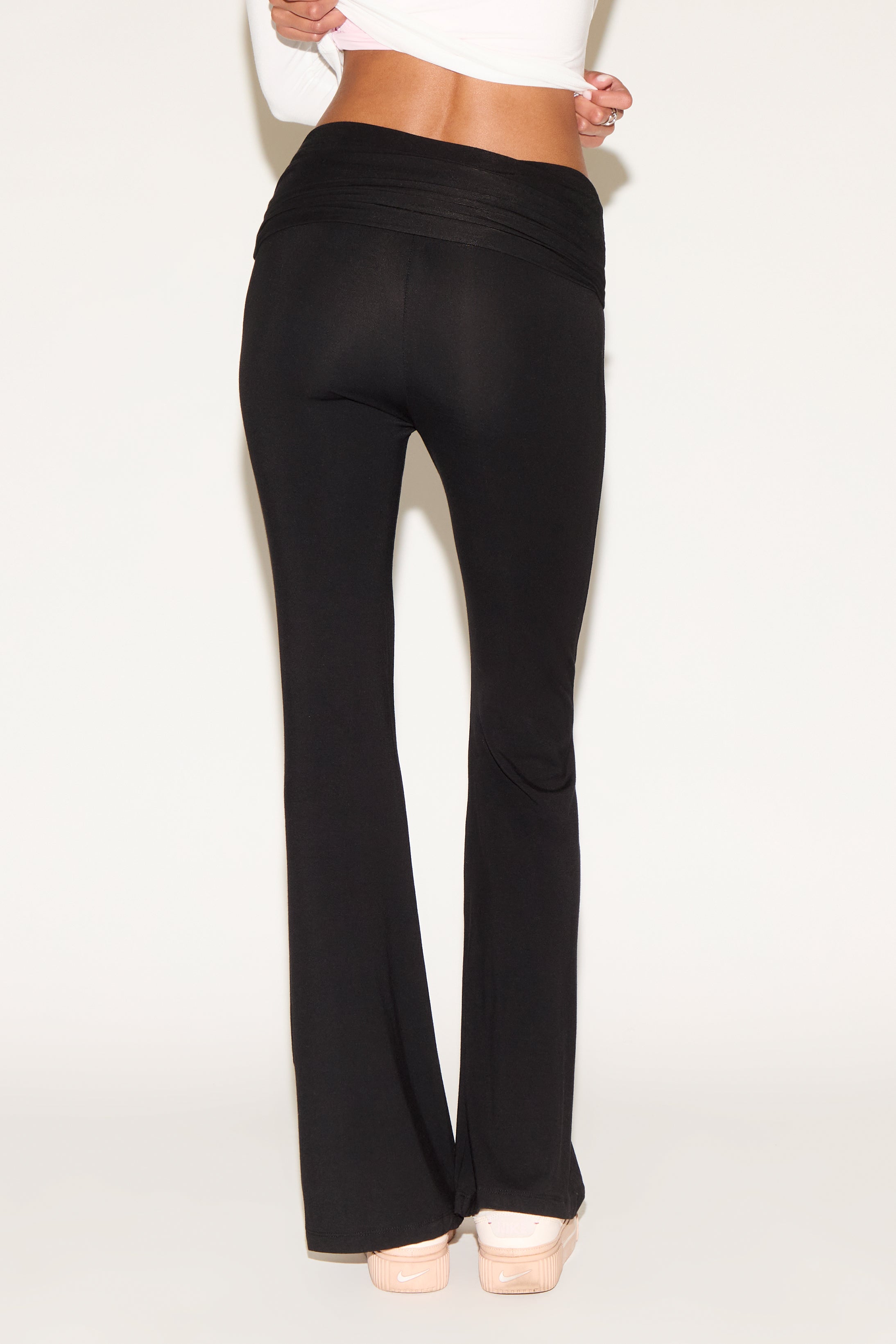 Model wearing black low-rise flare pants with a fold-over waistband, styled with a white long-sleeve top and white sneakers against a neutral background.
