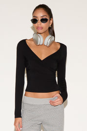 Model wearing a black long-sleeve wrap top with a V-neckline, styled with striped low-rise pants, silver over-ear headphones, and black oval sunglasses against a neutral background.