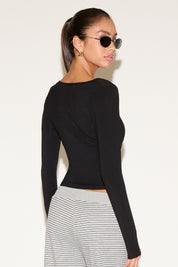 Back view of model wearing a black long-sleeve fitted wrap top, styled with striped low-rise pants and black oval sunglasses, hair pulled into a sleek ponytail against a neutral background.