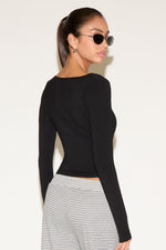 Back view of model wearing a black long-sleeve fitted wrap top, styled with striped low-rise pants and black oval sunglasses, hair pulled into a sleek ponytail against a neutral background.