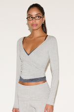 Model wearing a light gray long-sleeve wrap top with a deep V neckline and contrast trim, styled with matching low-rise pants and black rectangular glasses against a neutral background.