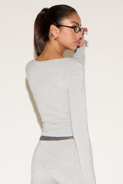 Back view of model wearing a light gray long-sleeve fitted wrap top, styled with matching low-rise pants and black rectangular glasses, hair in a sleek ponytail against a neutral background.