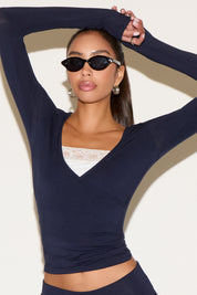 Model wearing a navy long-sleeve wrap top with a V-neckline, layered over a white lace bralette, styled with black oval sunglasses and silver hoop earrings against a neutral background.