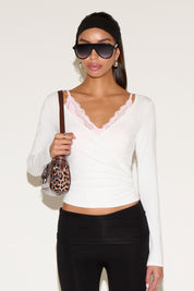 Model wearing a white long-sleeve wrap top with a V-neckline and lace trim, styled with black pants, oversized sunglasses, a black headband, and a leopard-print shoulder bag against a neutral background.