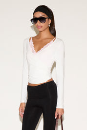 Model wearing a white long-sleeve wrap top with a V-neckline and lace trim, styled with black pants, oversized sunglasses, and a black headband, posed against a neutral background.