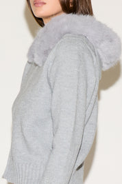 Close-up of a person wearing a gray knitted sweater with a fur-lined hood against a white background