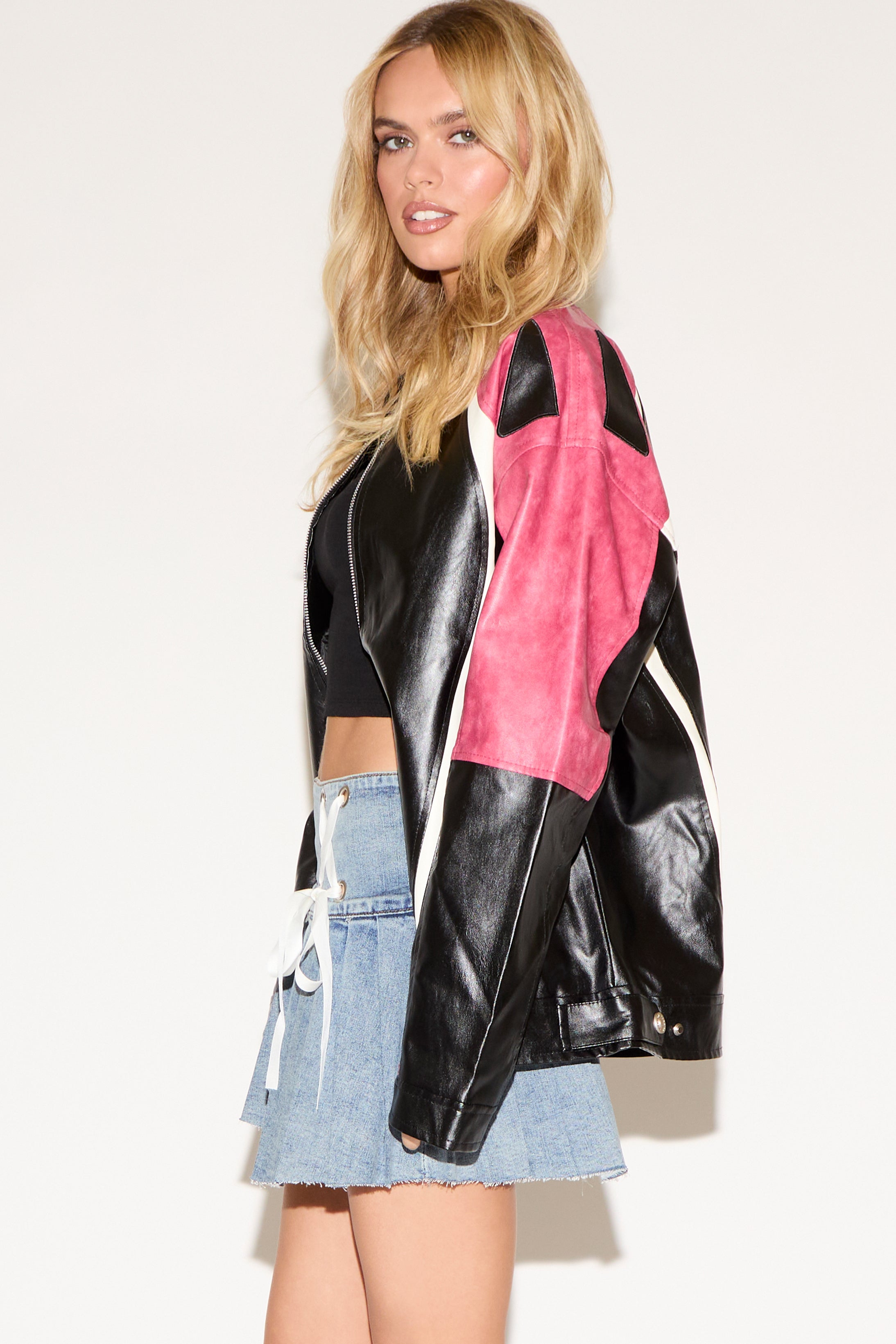 Woman wearing a black crop top, pink and black leather jacket, and a denim skirt with lace-up details. She poses confidently against a plain background.