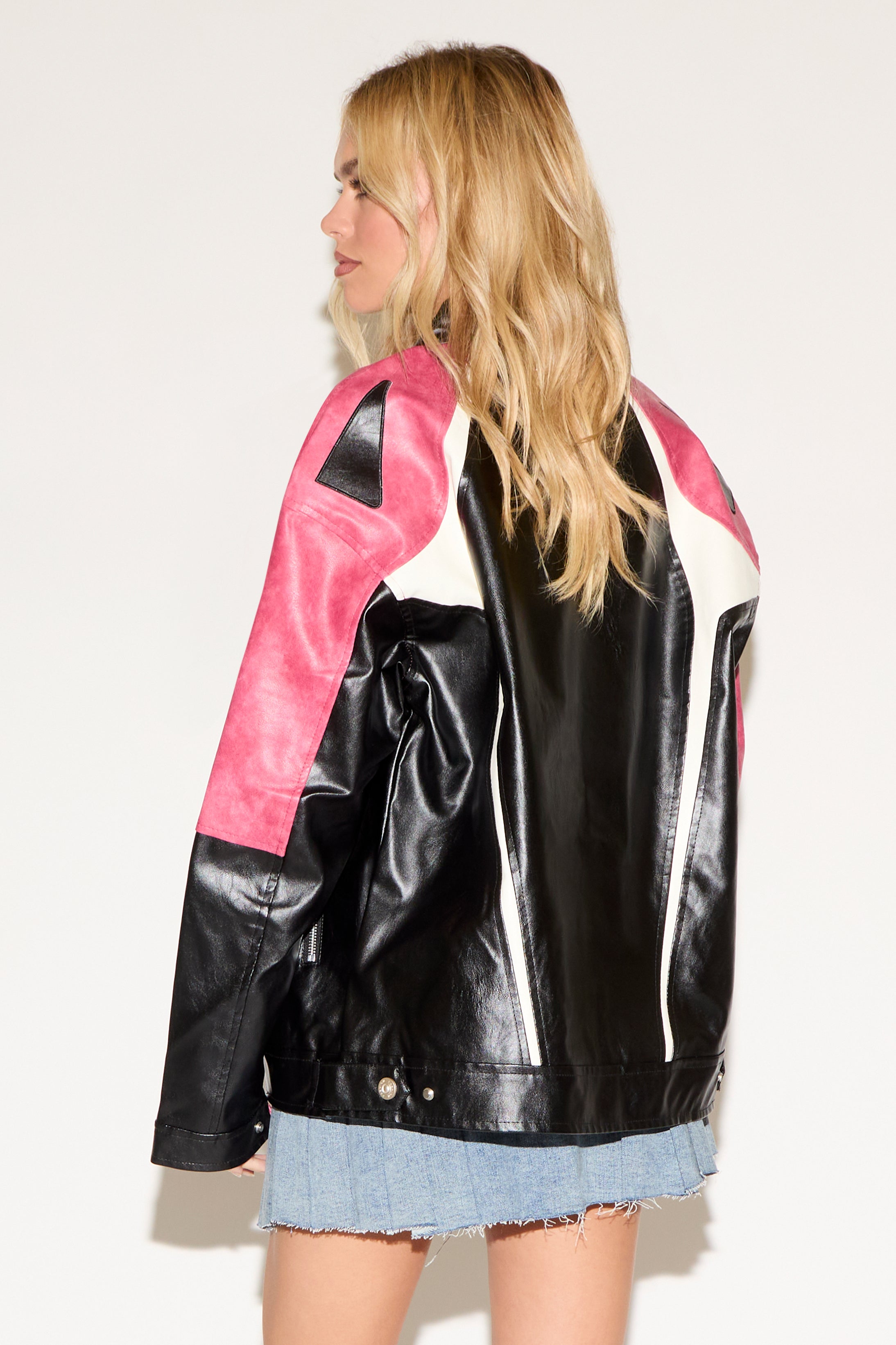 Woman wearing a black crop top, pink and black leather jacket, and a denim skirt with lace-up details. She poses confidently against a plain background.