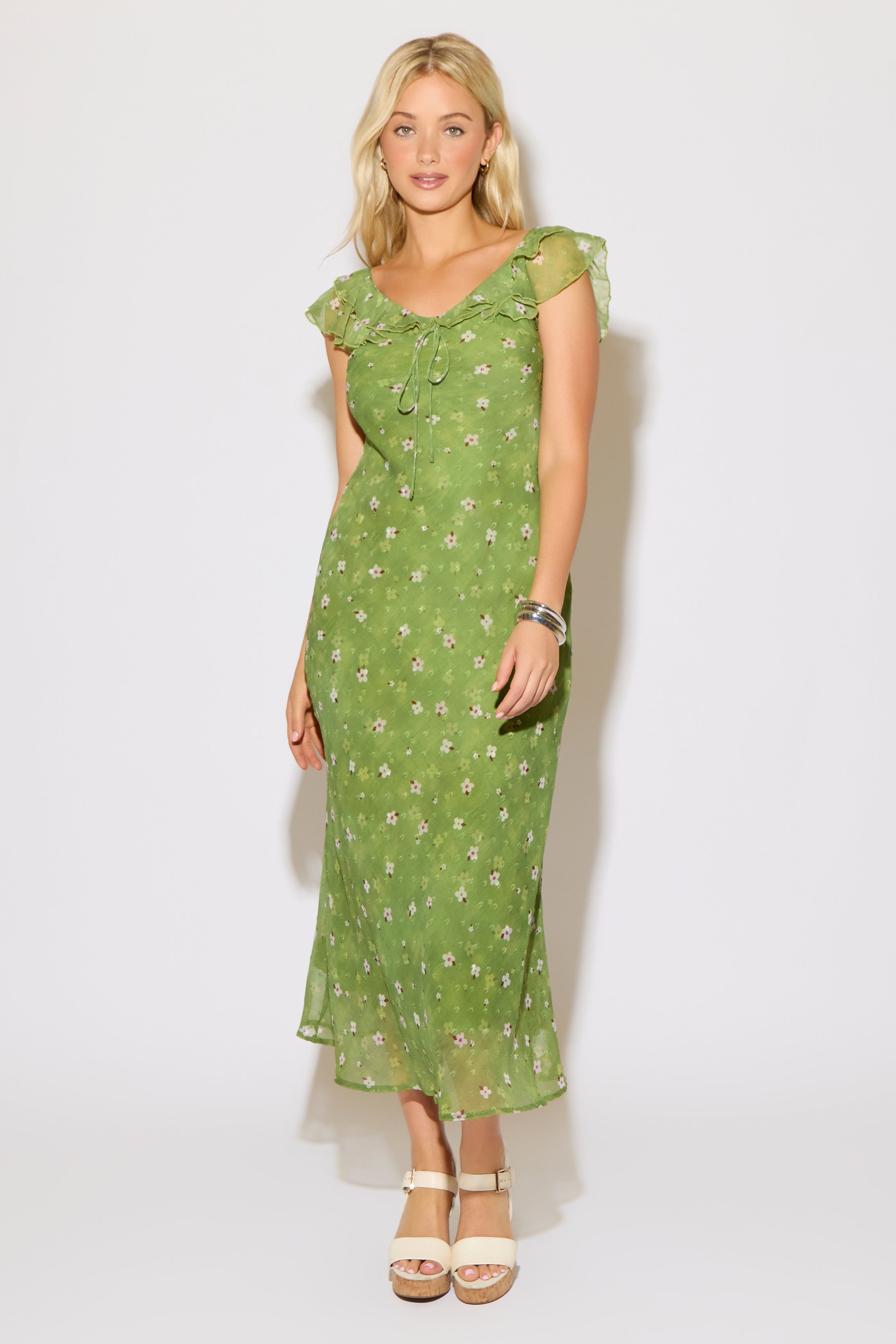 Model is wearing a green mesh floral overlay dress with ruffle detail on a white background. 
